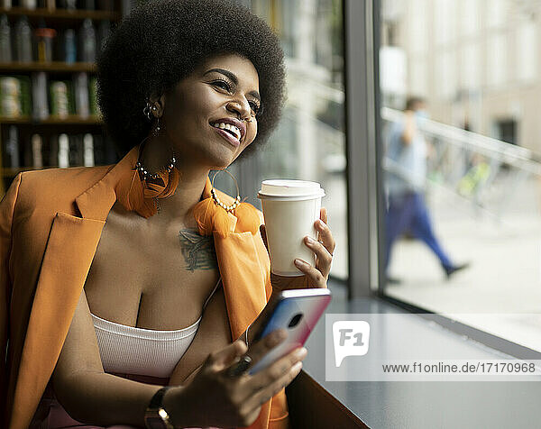 Happy woman with smart phone and coffee cup looking through window in cafe