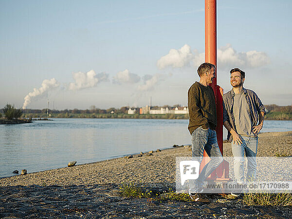 Smiling son and father leaning by pole while talking with each other at riverbank