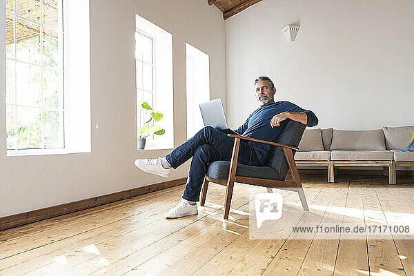 Älterer Geschäftsmann  der einen Laptop benutzt  während er zu Hause auf einem Sessel sitzt