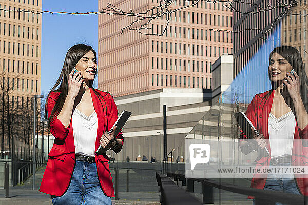 Smiling businesswoman with laptop talking on smart phone while looking at reflection