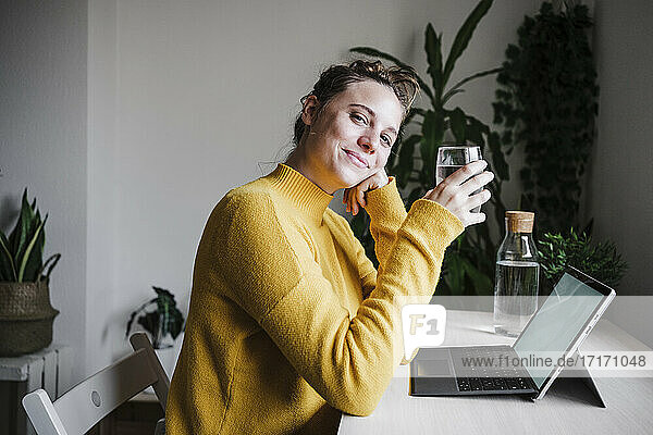Smiling woman drinking water while using digital tablet sitting at home office