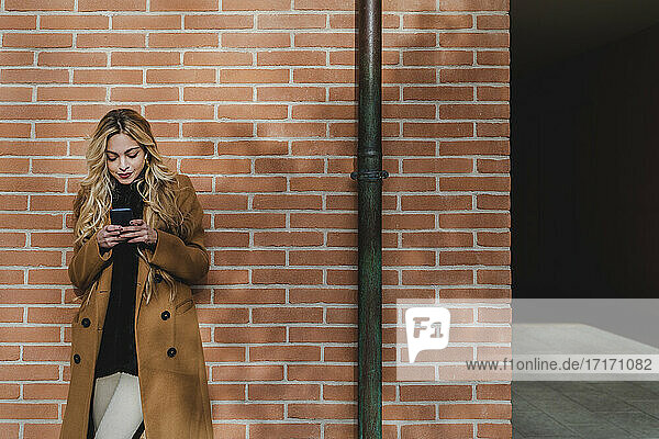 Young woman using smart phone while listening music against brick wall
