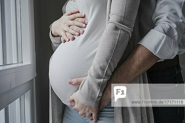 Schwangere Frau mit den Händen ihres Mannes auf dem Bauch  die zu Hause am Fenster steht