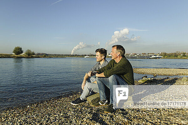 Father and son admiring river while sitting on rock during sunny day