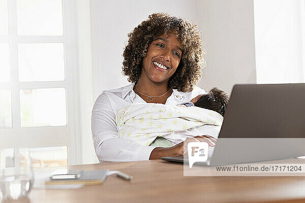 Smiling mother holding baby while working on laptop at home office