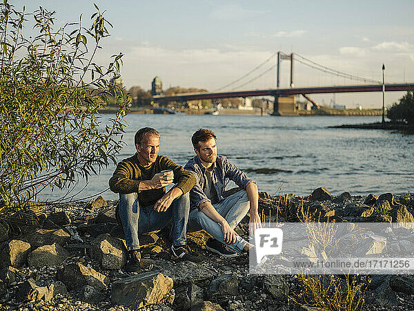Father and son sitting on rock against river at evening