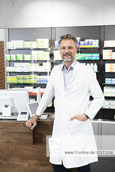 Smiling mature pharmacist with hand in pocket standing at checkout in pharmacy