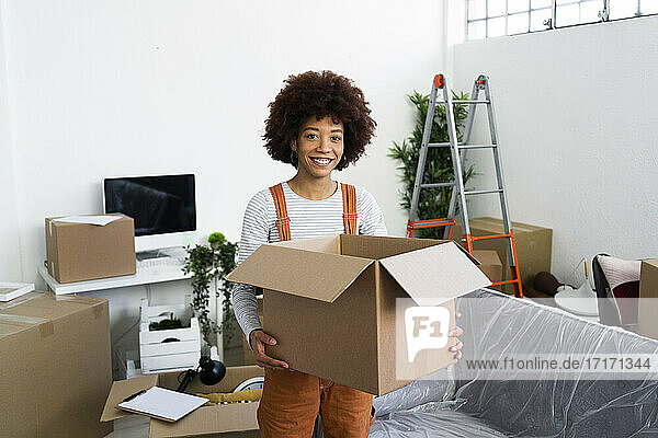 Lächelnder junger Mann  der einen Karton in der Hand hält  während er bei einem Umzug in seinem neuen Zuhause steht