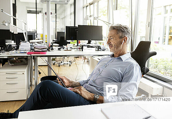 Geschäftsmann benutzt ein digitales Tablet  während er sich auf einem Stuhl in einem Großraumbüro entspannt