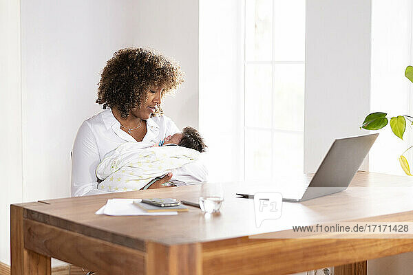 Mutter sieht schlafendes Baby an  während sie am Tisch im Büro zu Hause sitzt