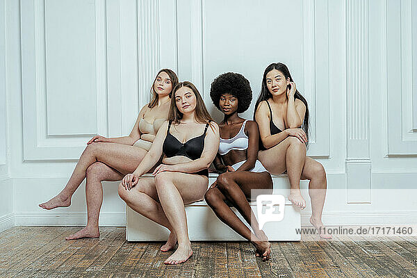 Confident multi-ethnic group of models in lingerie sitting against white wall