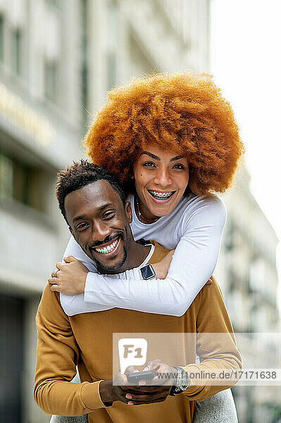 Smiling woman piggybacking on man using mobile phone while standing outdoors