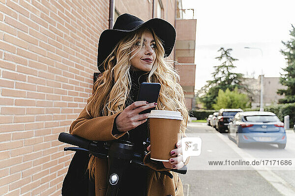 Young woman with electric push scooter holding reusable coffee cup while looking away