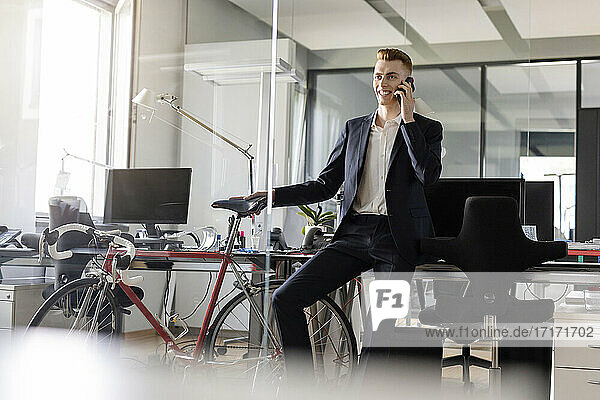 Male entrepreneur with bicycle smiling while talking on mobile phone sitting at open plan office