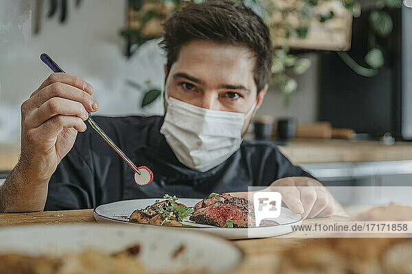 Male chef wearing protective face mask garnishing vegetable on tomahawk steak while working in kitchen