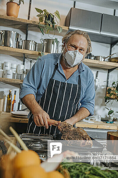Mature expertise wearing face mask grilling tomahawk steak in frying pan while standing in kitchen