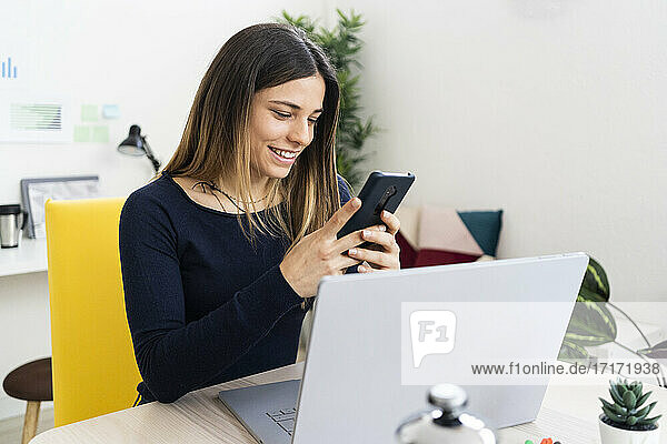 Lächelnder Freiberufler  der ein Mobiltelefon benutzt  während er im Wohnzimmer am Laptop sitzt