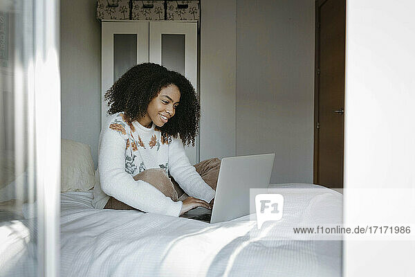 Lächelnde Frau  die einen Laptop benutzt  während sie zu Hause auf dem Bett sitzt