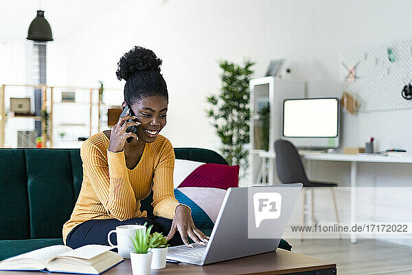 Smiling young woman talking on mobile phone while using laptop in living room