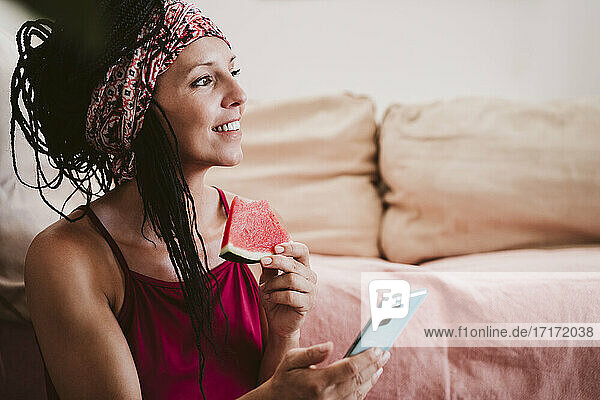 Frau mit Handy isst Wassermelone  während sie zu Hause sitzt