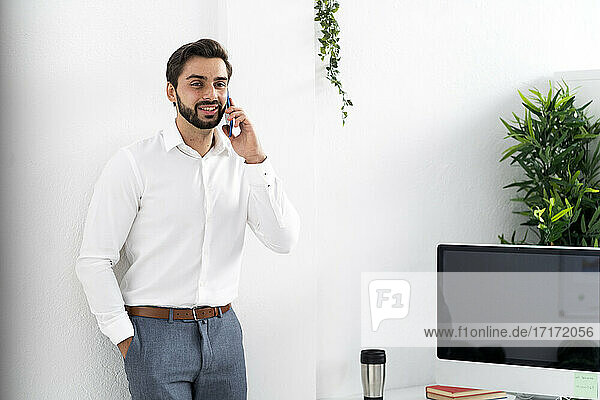 Männlicher Berufstätiger mit Händen in den Taschen bei einem Telefongespräch an einer weißen Wand am Arbeitsplatz