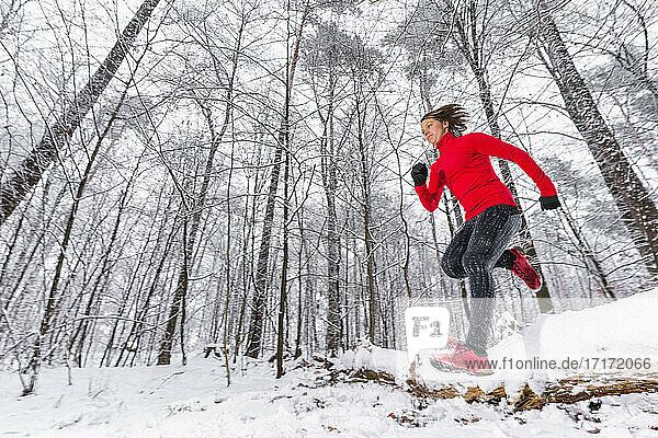 Young female athlete jumping over snow covered fallen tree in forest