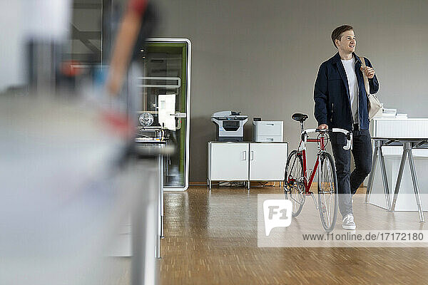 Businessman looking away while walking with bicycle at open plan office