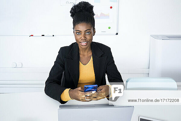 Young female professional text messaging through mobile phone while sitting with laptop at desk