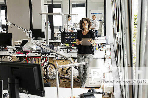 Geschäftsfrau arbeitet an einem digitalen Tablet  während sie mit einem Kollegen im Hintergrund in einem Großraumbüro steht
