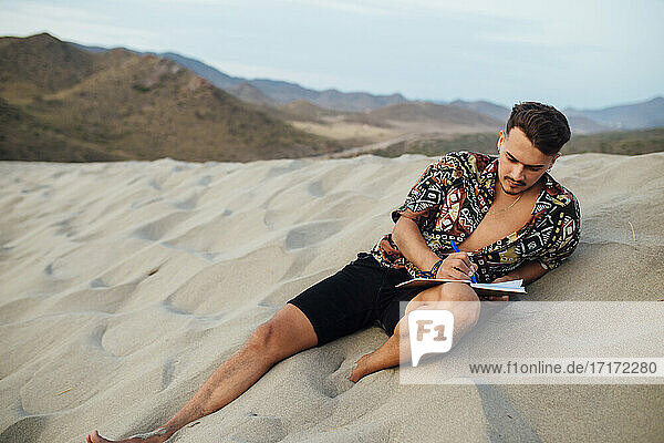 Gut aussehender Mann  der in ein Buch schreibt  während er auf dem Sand in Almeria  Tabernas-Wüste  Spanien  sitzt