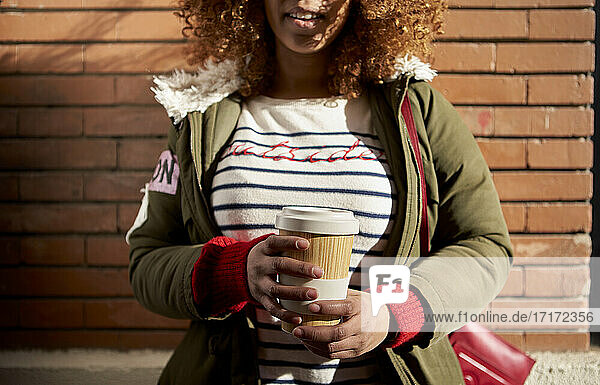 Woman wearing jacket holding disposable coffee cup while standing against brick wall