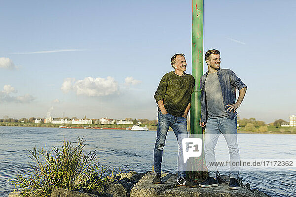 Smiling son and father leaning by pole while looking away at riverbank against sky