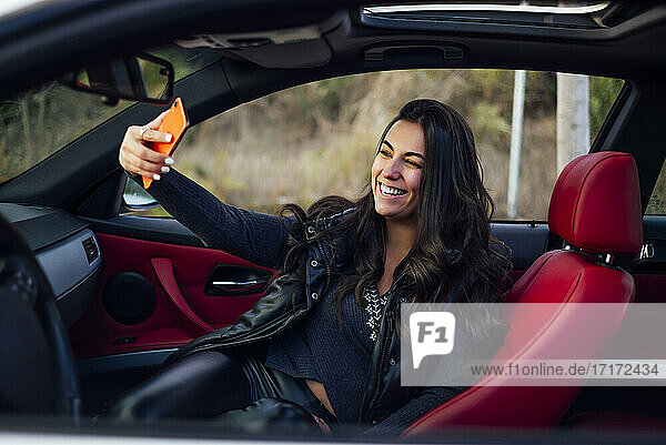 Smiling woman taking selfie through smart phone while sitting in car