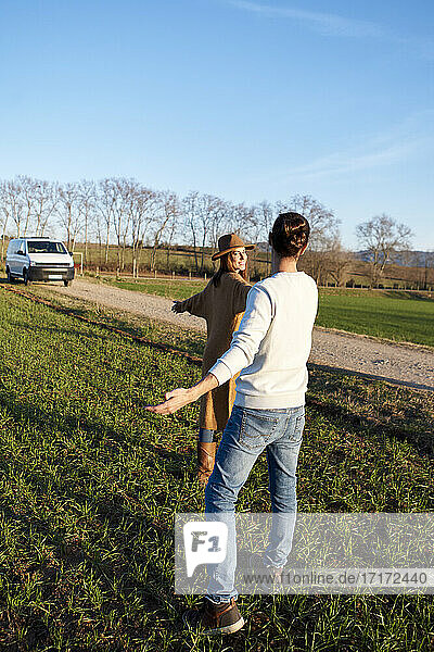 Heterosexual couple with arms outstretched standing on green land during sunset