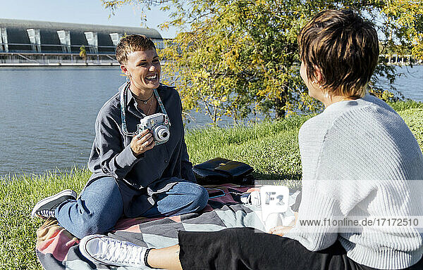 Smiling woman with camera looking at friend while sitting against river at park