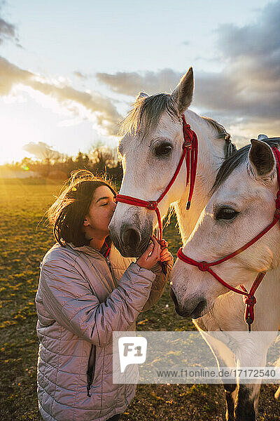 Girl wearing jacket kissing horses while standing in ranch