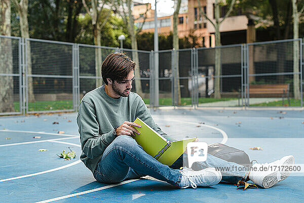 Junger Mann studiert beim Lesen eines Buches auf einem Basketballplatz in der Universität