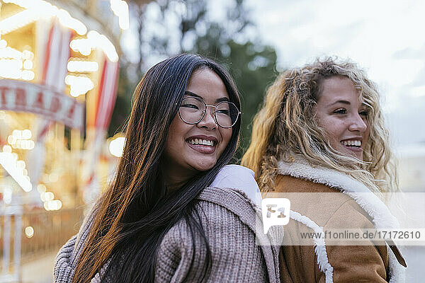 Multi-ethnic female friends looking away while standing back to back by illuminated carousel