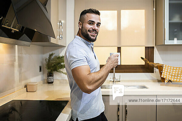 Lächelnder muskulöser Mann  der eine Kaffeetasse hält  während er sich zu Hause an den Küchentisch lehnt