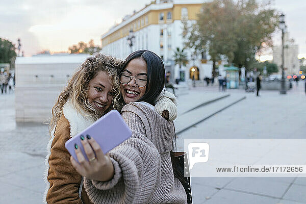 Cheerful female friends taking selfie through smart phone while embracing on sidewalk in city