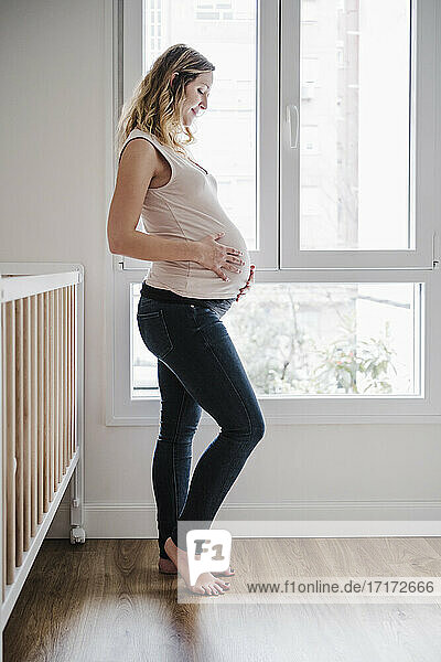 Schwangere Mutter mit Händen auf dem Bauch in Erwartung am Fenster stehend