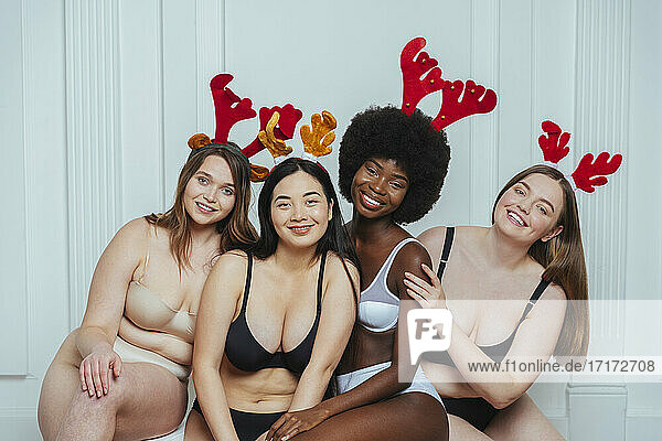 Lächelnde multiethnische Gruppe von Model-Freundinnen in Unterwäsche mit Rentiergeweih vor weißer Wand