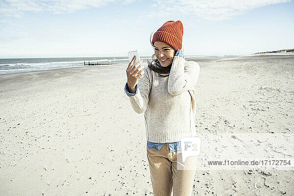 Porträt einer Frau mit Strickmütze  die am Strand mit ihrem Smartphone telefoniert
