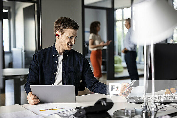 Young businessman using mobile phone while sitting with colleague in background at open plan office