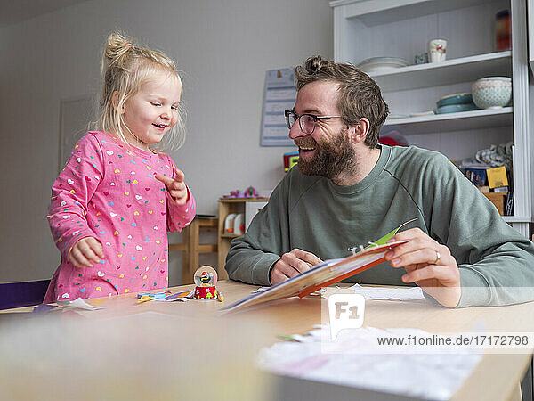 Happy father showing book to cute daughter on table at home