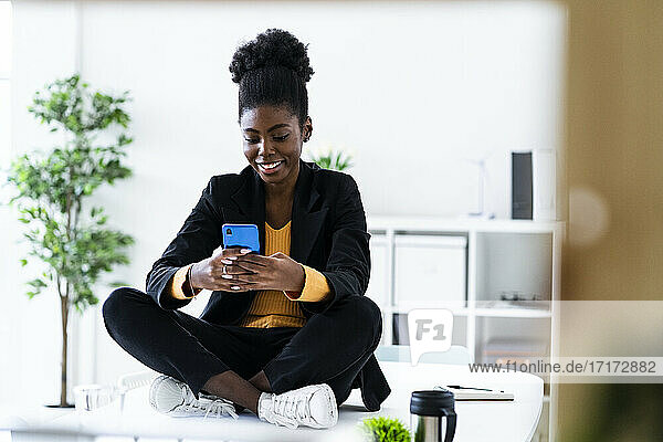 Smiling young Afro female entrepreneur text messaging using smart phone while sitting cross-legged on desk