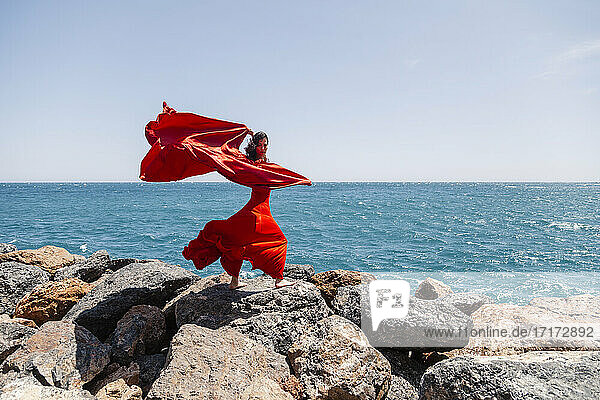 Mature female dancer in red dress dancing with sarong on rocks by seashore