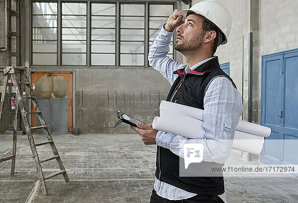 Männlicher Architekt mit digitalem Tablet und Bauplänen  der in einem Gebäude steht und nach oben schaut