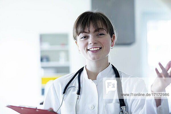 Smiling female doctor gesturing while holding clipboard in hospital