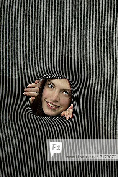 Portrait of young woman smiling through hole in gray striped background